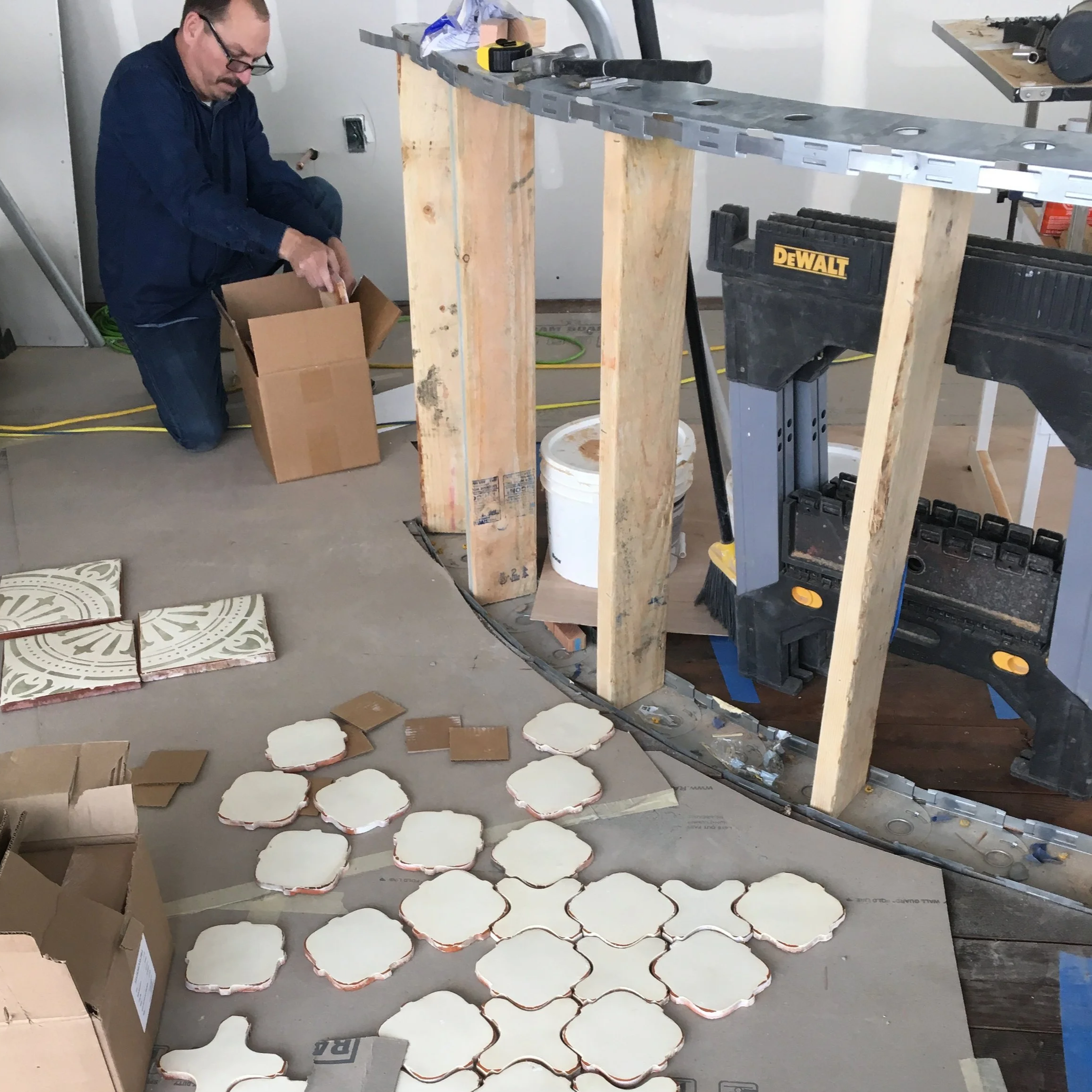 A man working on a construction project, assembling or organizing decorative tiles with a floral design on the floor in a room with unfinished walls and various tools and materials around.