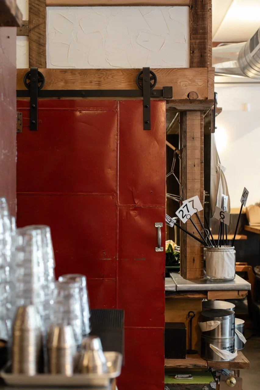 Part of a bar or cafe interior showing a red metal door, wooden beams, a jar with black sticks labeled with numbers, and glassware in the foreground.