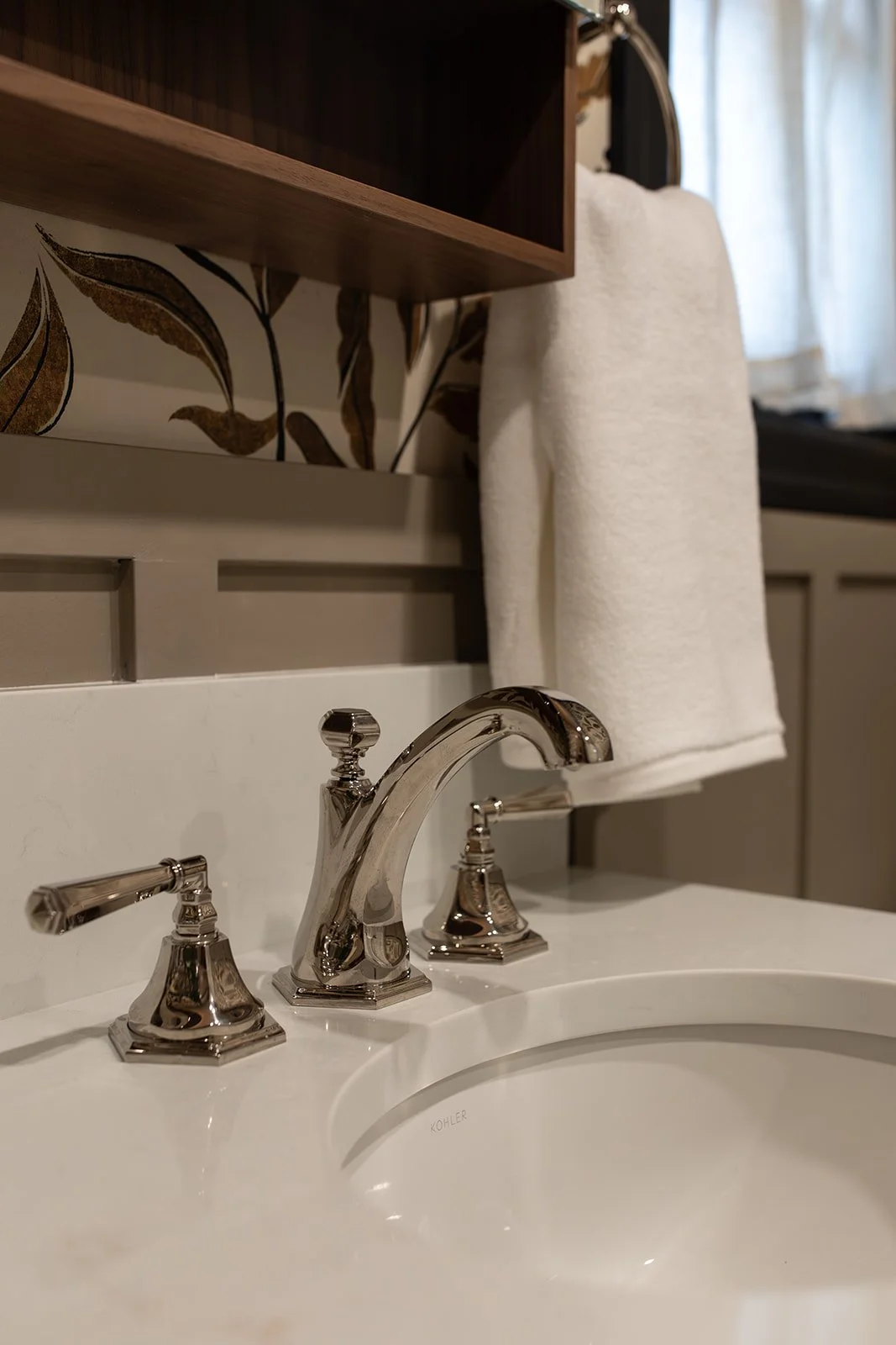 Close-up of a cream-colored bathroom sink with a shiny silver faucet and handles, a white towel hanging on a towel rack, and a wooden shelf above with a leaf-patterned wallpaper in the background.