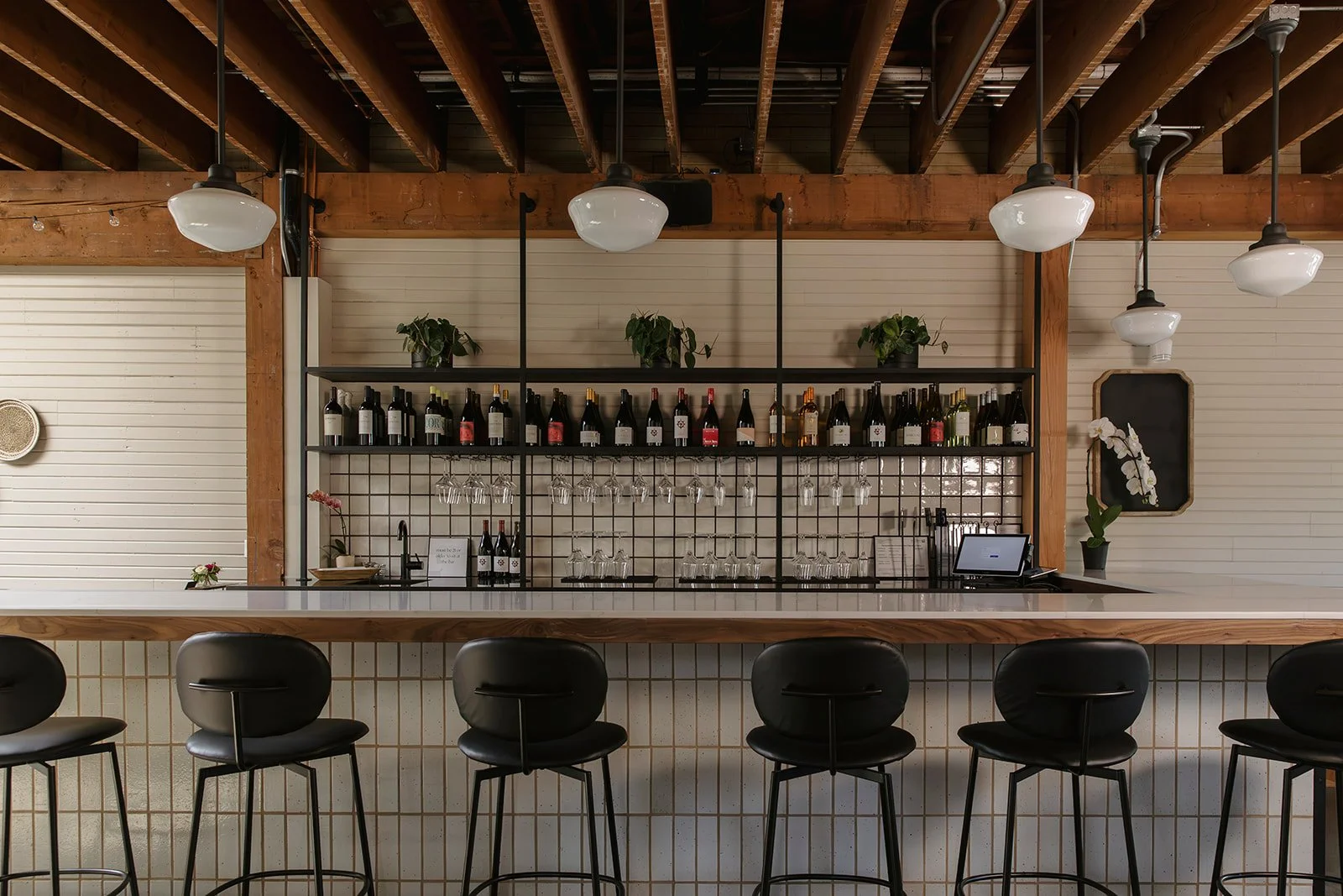 Modern bar with black chairs, a white countertop, shelves with wine bottles and glasses, decorated with green plants and orchids, wooden beams and pendant lights overhead.