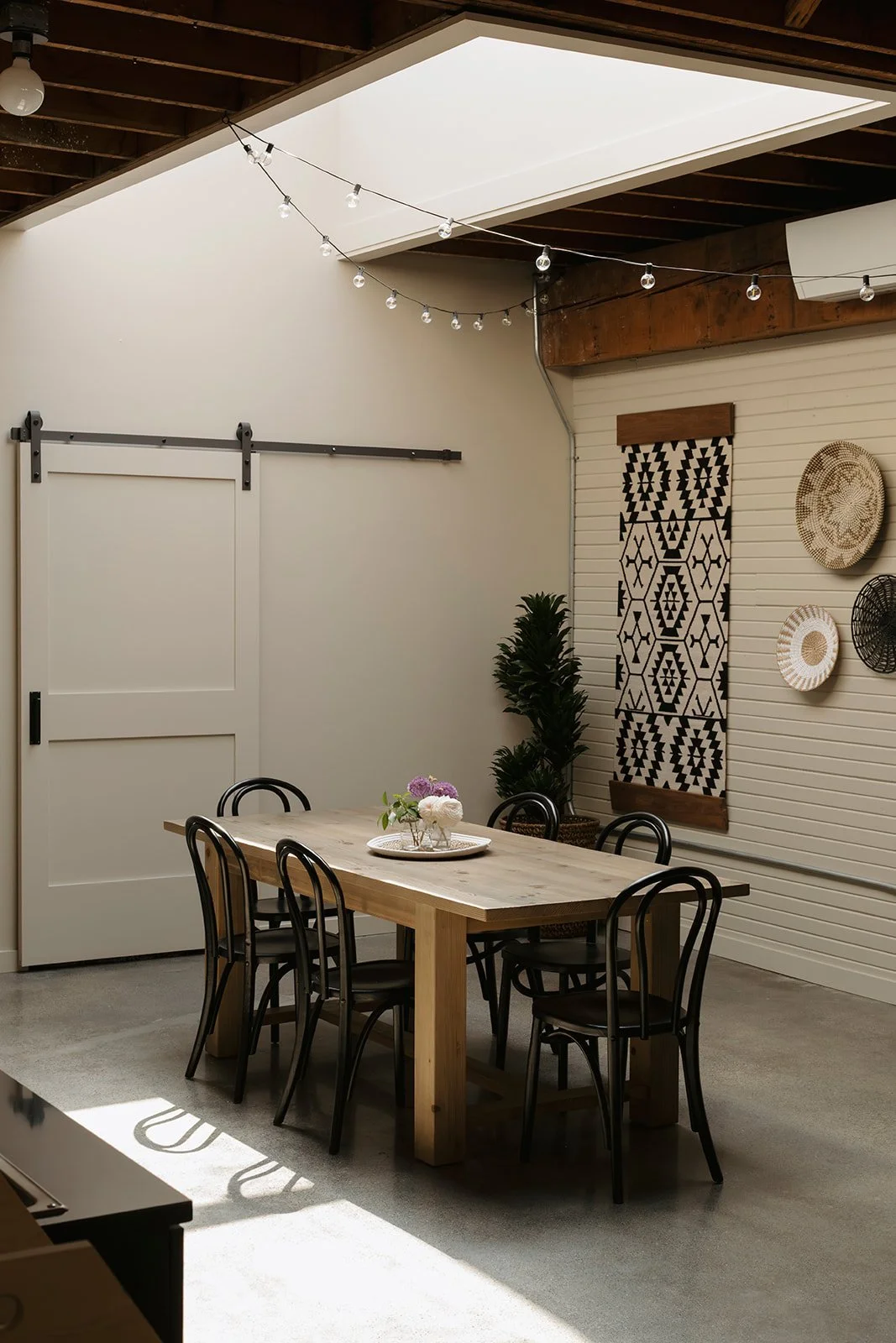 A dining room with a wooden table and six black chairs, decorative plates on the wall, fringe wall hanging, string lights overhead, and a sliding barn door.