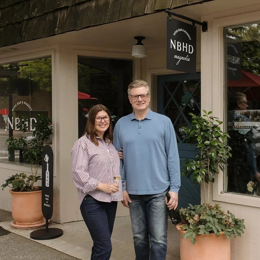 A man and woman standing outside a wine bar called NBHD Magnolia, smiling, with the woman holding a glass of white wine.