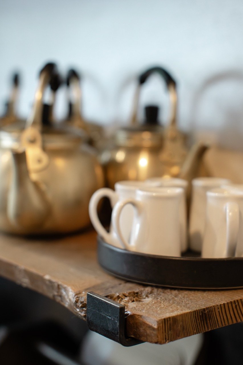 A wooden table holding two stainless steel teapots and several white ceramic mugs in a black tray, with a plain light-colored wall in the background.