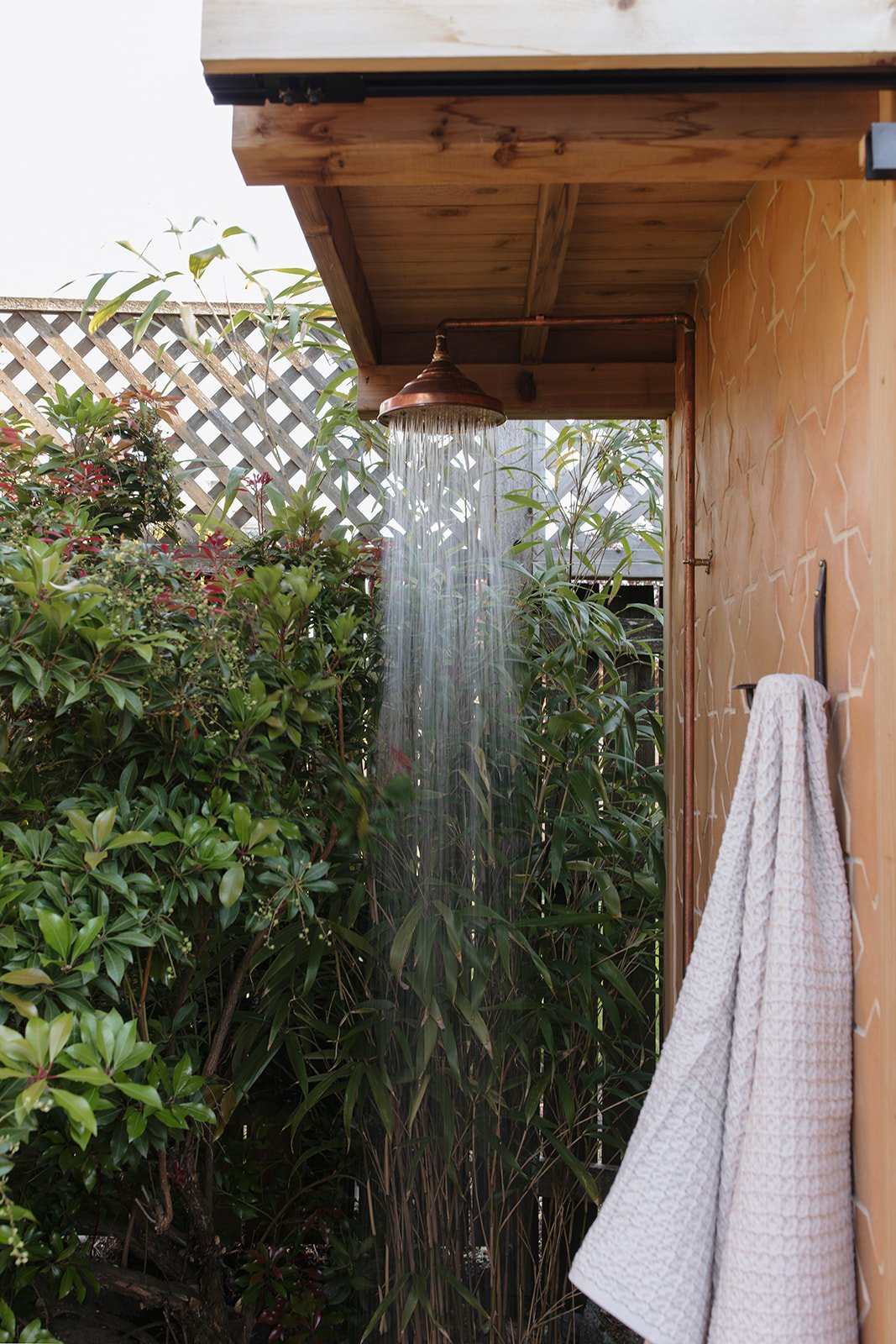 Outdoor shower with copper shower head and pipe, surrounded by green plants and a wooden lattice fence, with a towel hanging on a wall.