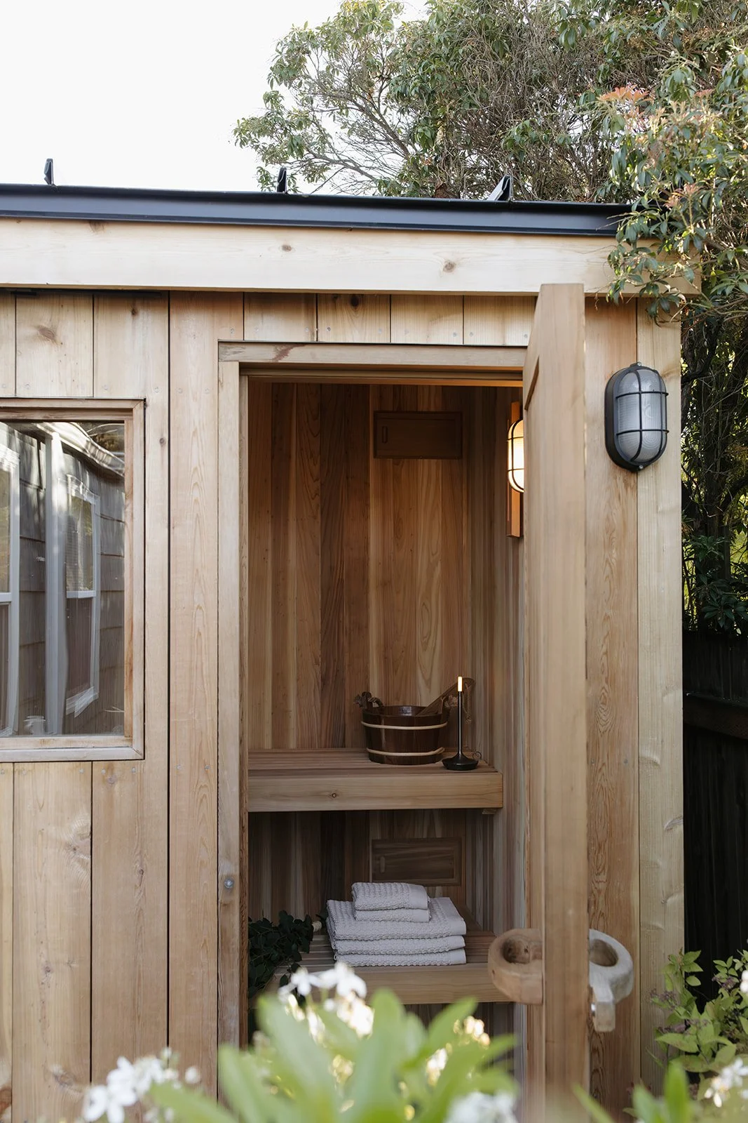 Small wooden outdoor saunas with a window, towels, sauna bucket, and a candle inside, surrounded by greenery.