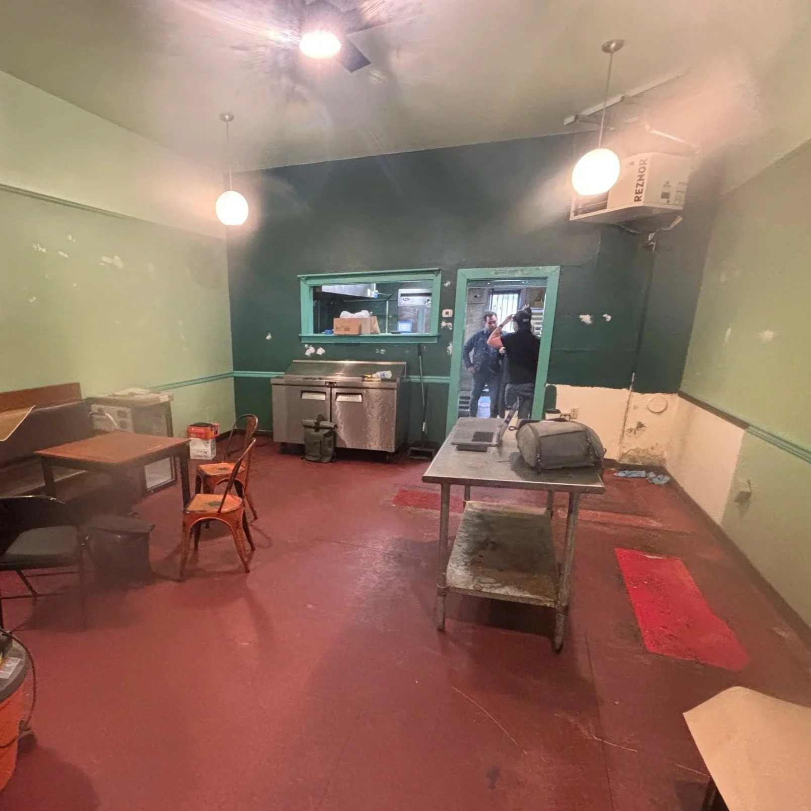 Empty dining area with tables and chairs, some furniture and boxes, a window with a teal border, a large stainless steel sink, and two hanging light fixtures. Two people are visible through a doorway in the background.