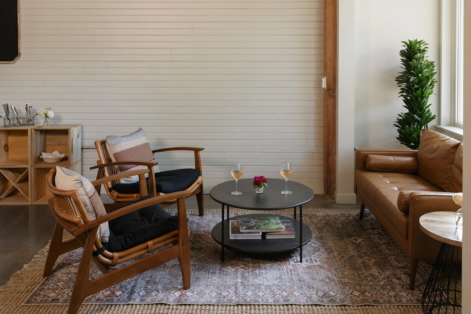 Cozy living room with wooden chairs, a leather sofa, a black coffee table with wine glasses and a flower arrangement, a patterned rug, a white wall, and a large green plant near the window.