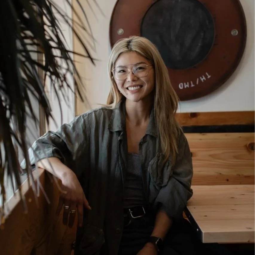 A woman with glasses and long hair smiling, sitting in a cozy room with wooden furniture and a large decorative circular piece on the wall behind her.