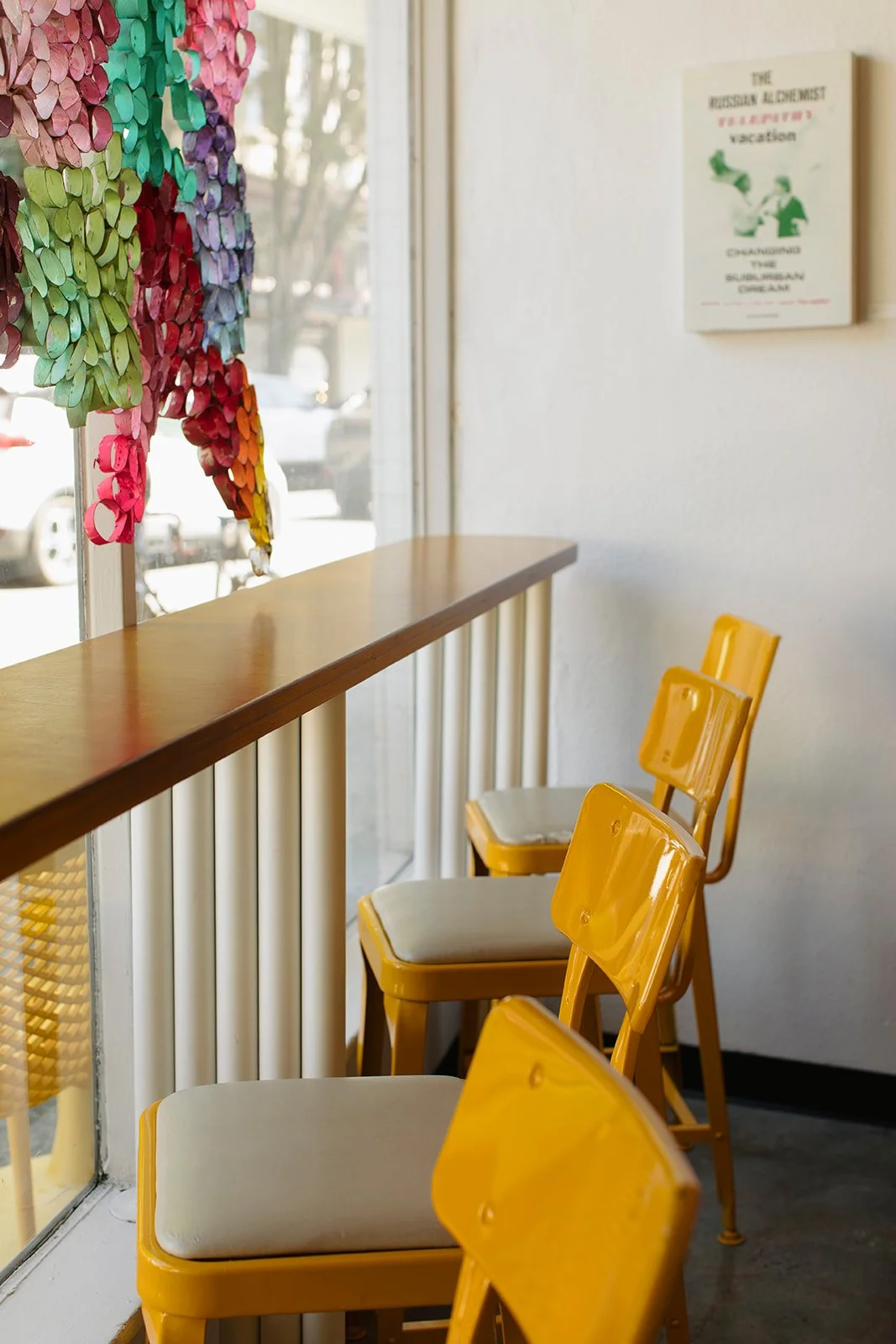 Inside a cafe, yellow chairs are lined up along a wooden counter next to large windows. Colorful paper or fabric decorations hang on the window, and a sign about a Russian alchemist is on the wall.
