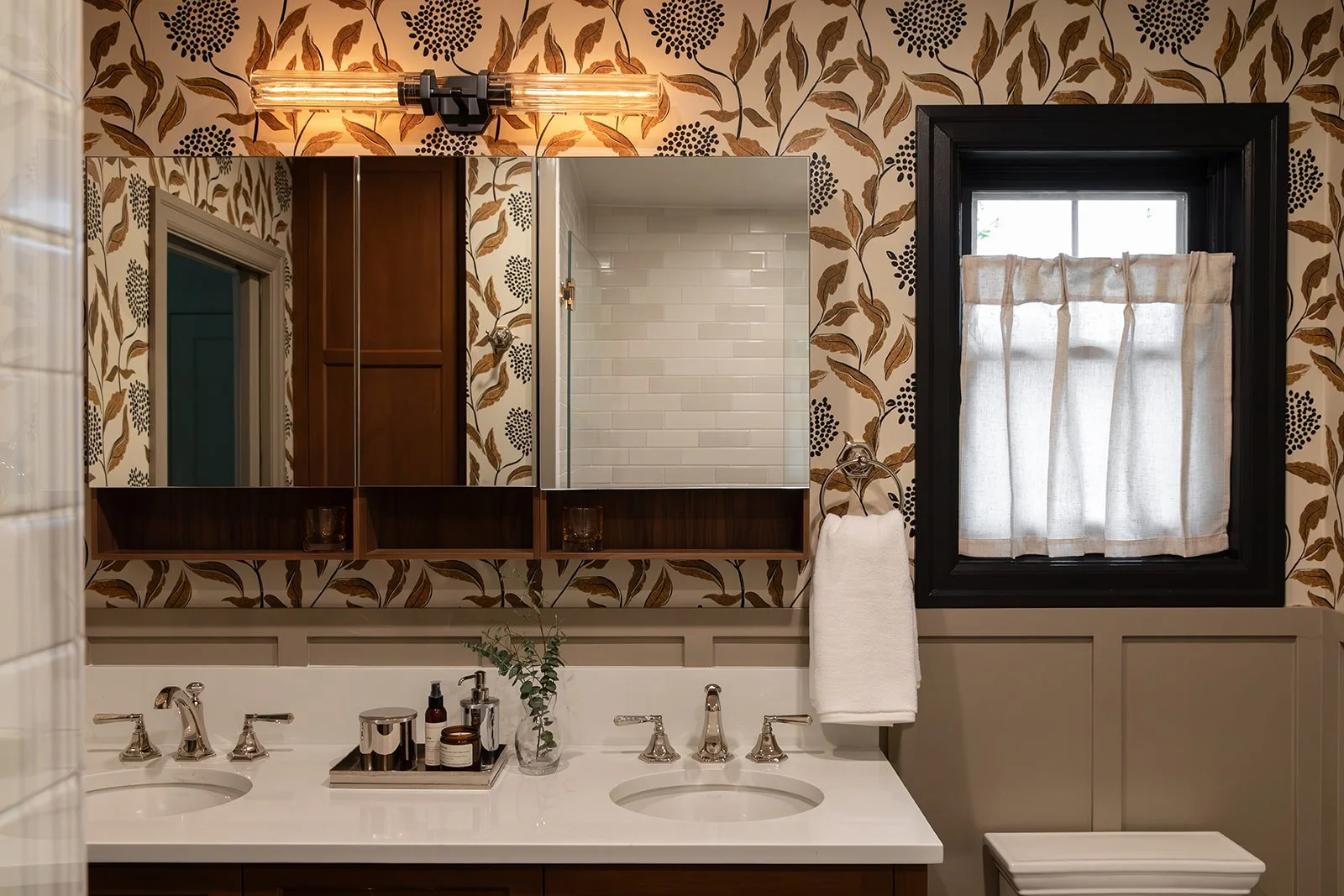 A bathroom with a double sink vanity, a mirror, and a window with curtains. The wallpaper has a floral pattern with dark berries and brown leaves. There is a light fixture above the mirror, a towel hanging on a ring, and a small potted plant on the counter.