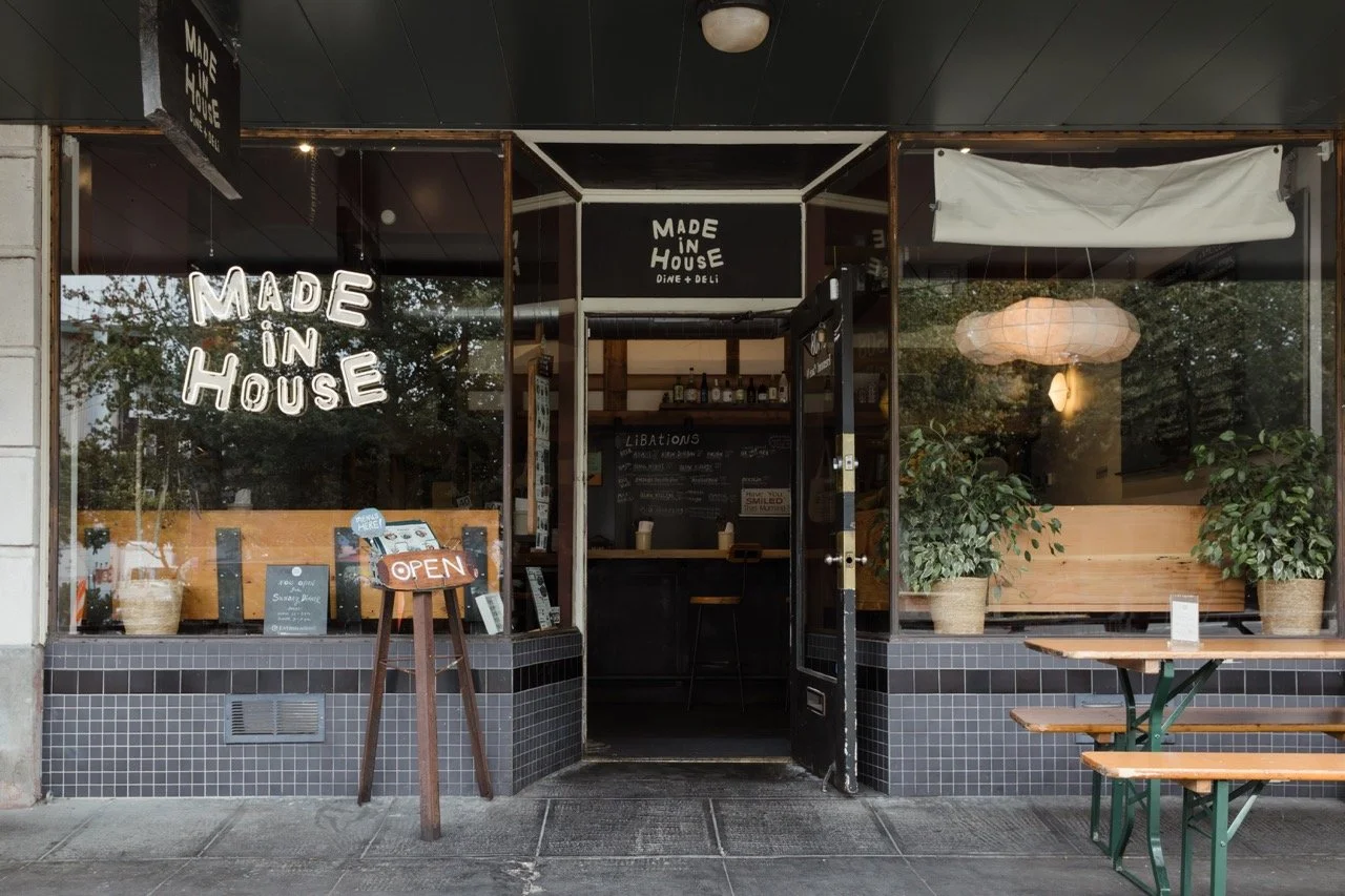 Exterior of a cafe with large glass windows displaying plants inside, white neon sign that says "Made in House", and an “Open” sign on a wooden stand outside the door.