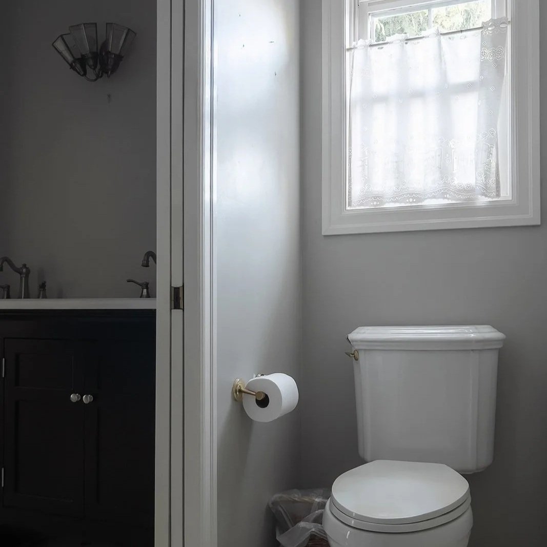 A small bathroom with a white toilet, a window with lace curtains, and a partial view of a dark vanity with a sink and faucet.