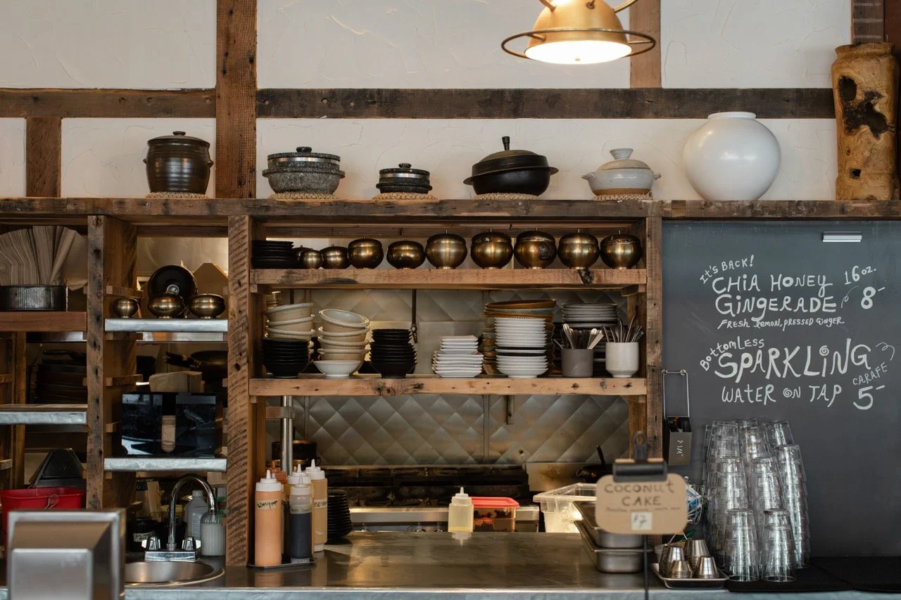 A rustic cafe kitchen with wooden shelves holding various ceramic and metal bowls, cups, and containers, and a black chalkboard menu with handwritten items featuring chai honey ginger tea and sparkling water.