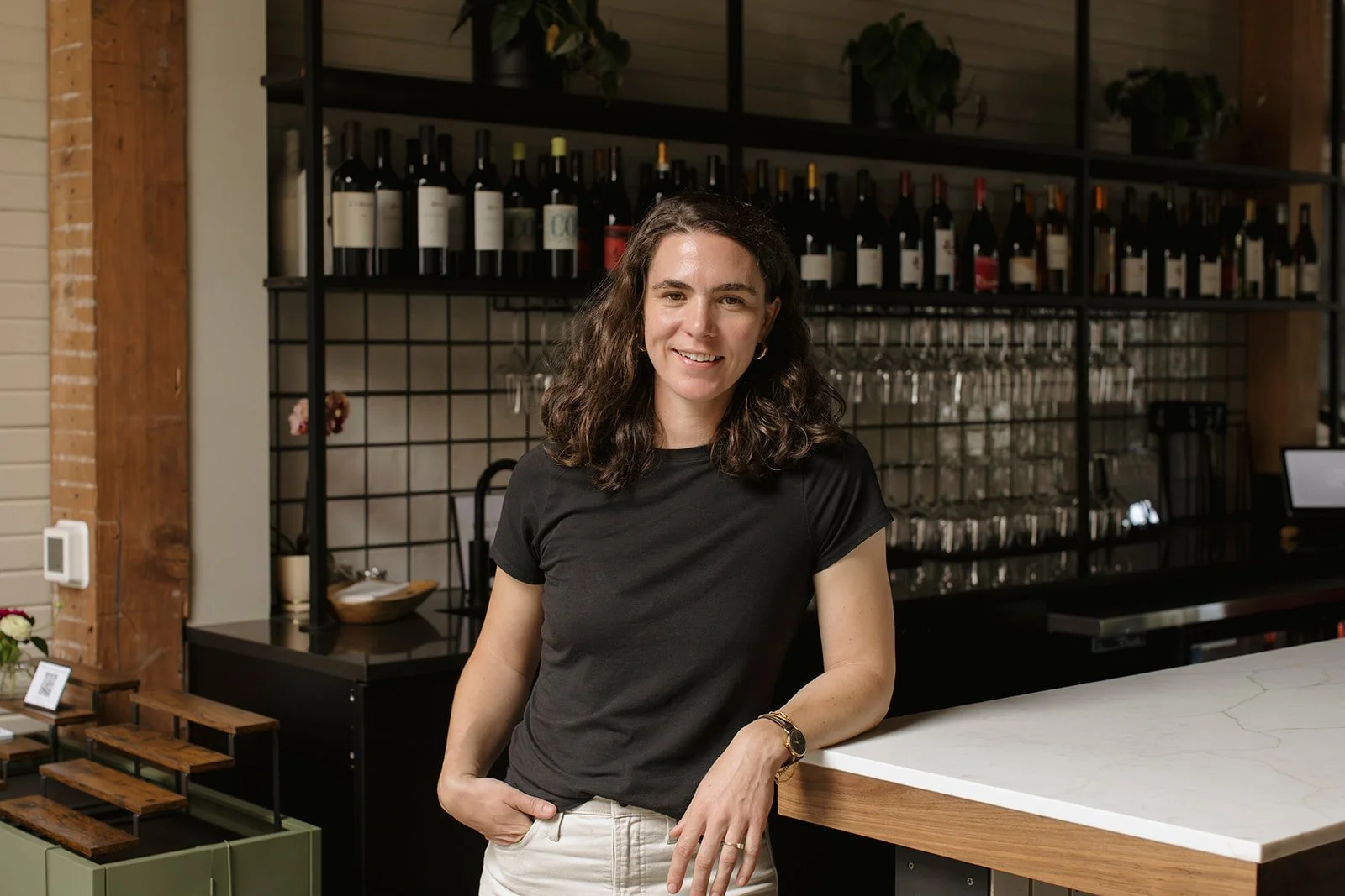 A woman standing behind a bar counter in a modern, stylish establishment with wine bottles and glasses in the background.