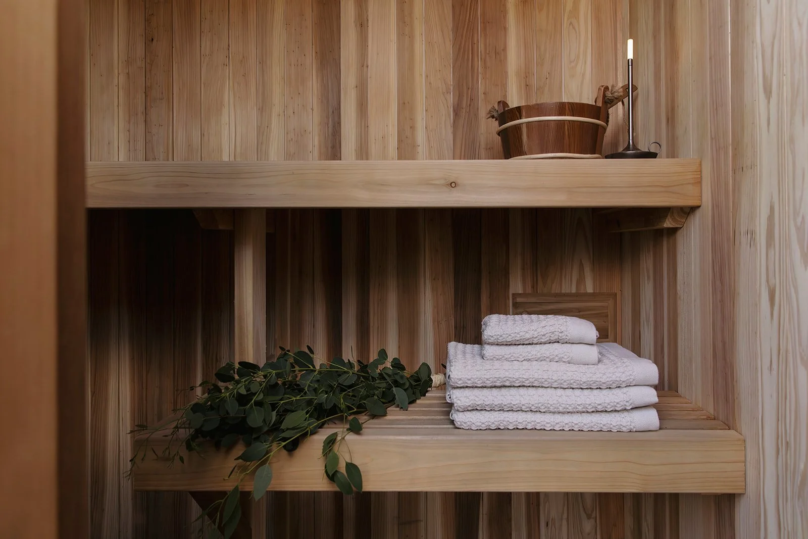 Wooden shelves inside a sauna with rolled white towels, green foliage, a wooden bucket, and a sauna light.