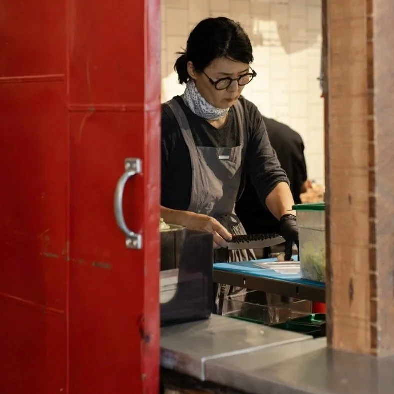 A woman with glasses, wearing a gray apron and a scarf, working in a kitchen or food preparation area, seen through a partially open red door.