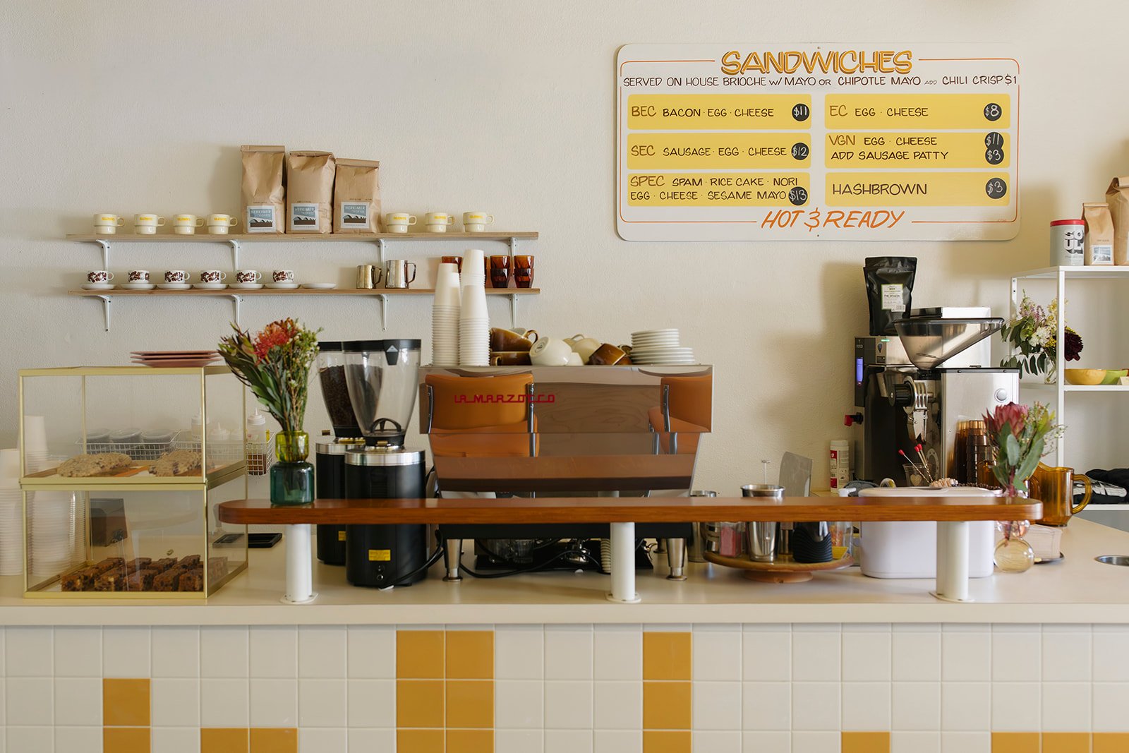 A small cafe counter with a coffee machine, glass jars, flowers, and baked goods in a display case, with a whiteboard menu listing sandwiches like bacon, egg, cheese, sausage, and hashbrown.