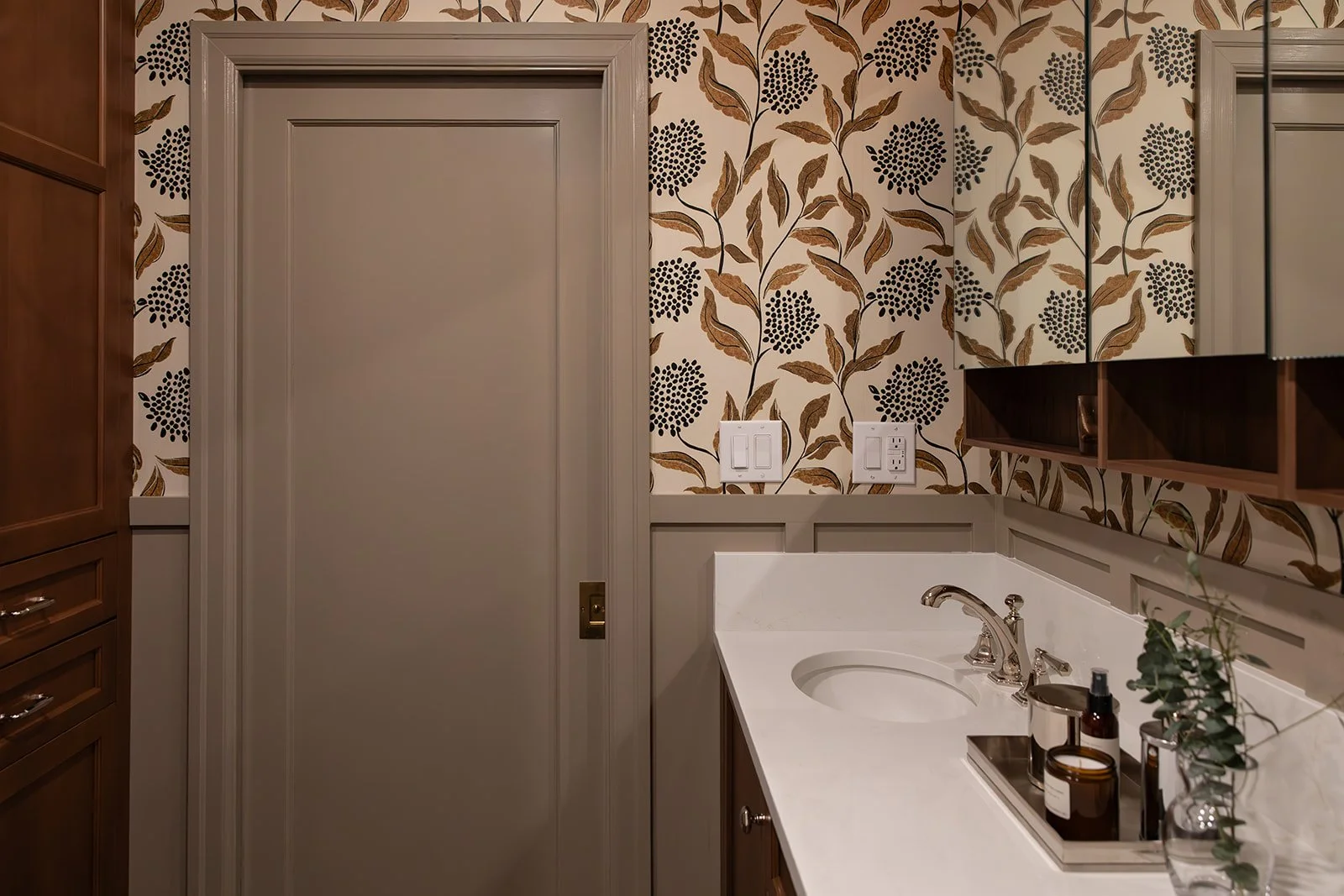 Bathroom vanity with a white countertop, sink, and chrome faucet, bathroom cabinets, floral patterned wallpaper with brown and black leaves, electrical outlets, and a mirror.