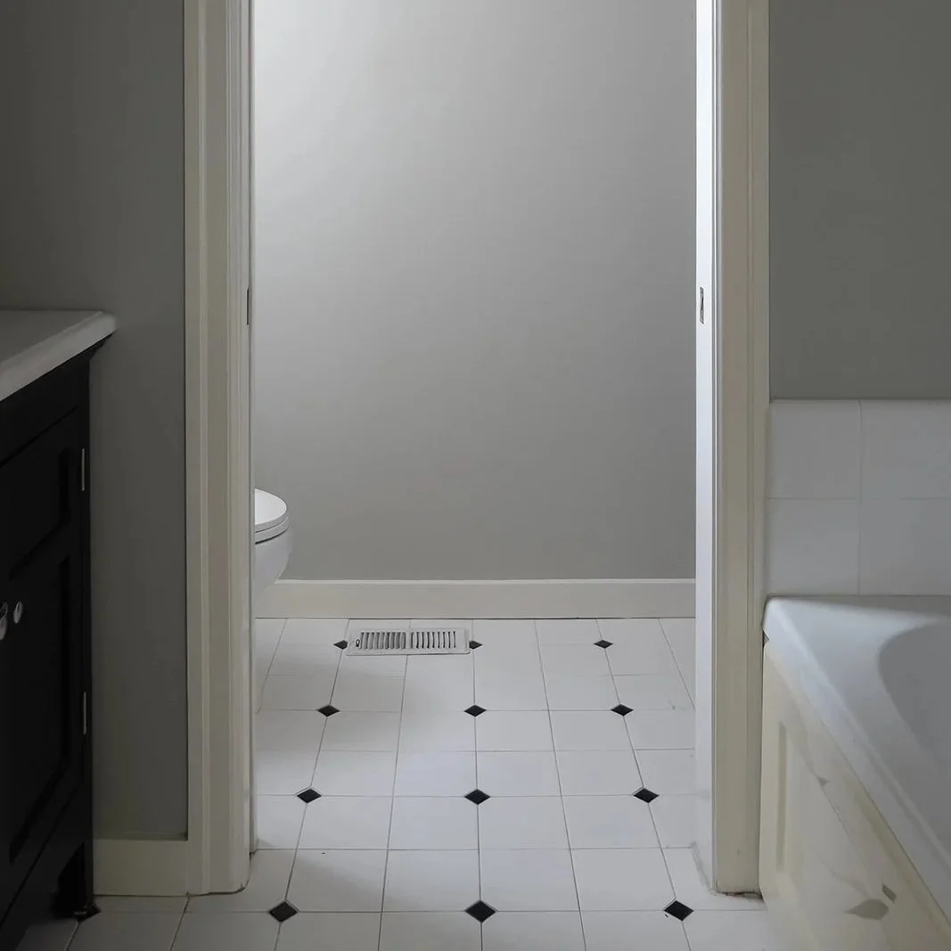 Bathroom with white tiled floor, black diamond accents, a vent on the floor, toilet partially visible on the left, and a bathtub on the right, with a gray wall in the background.
