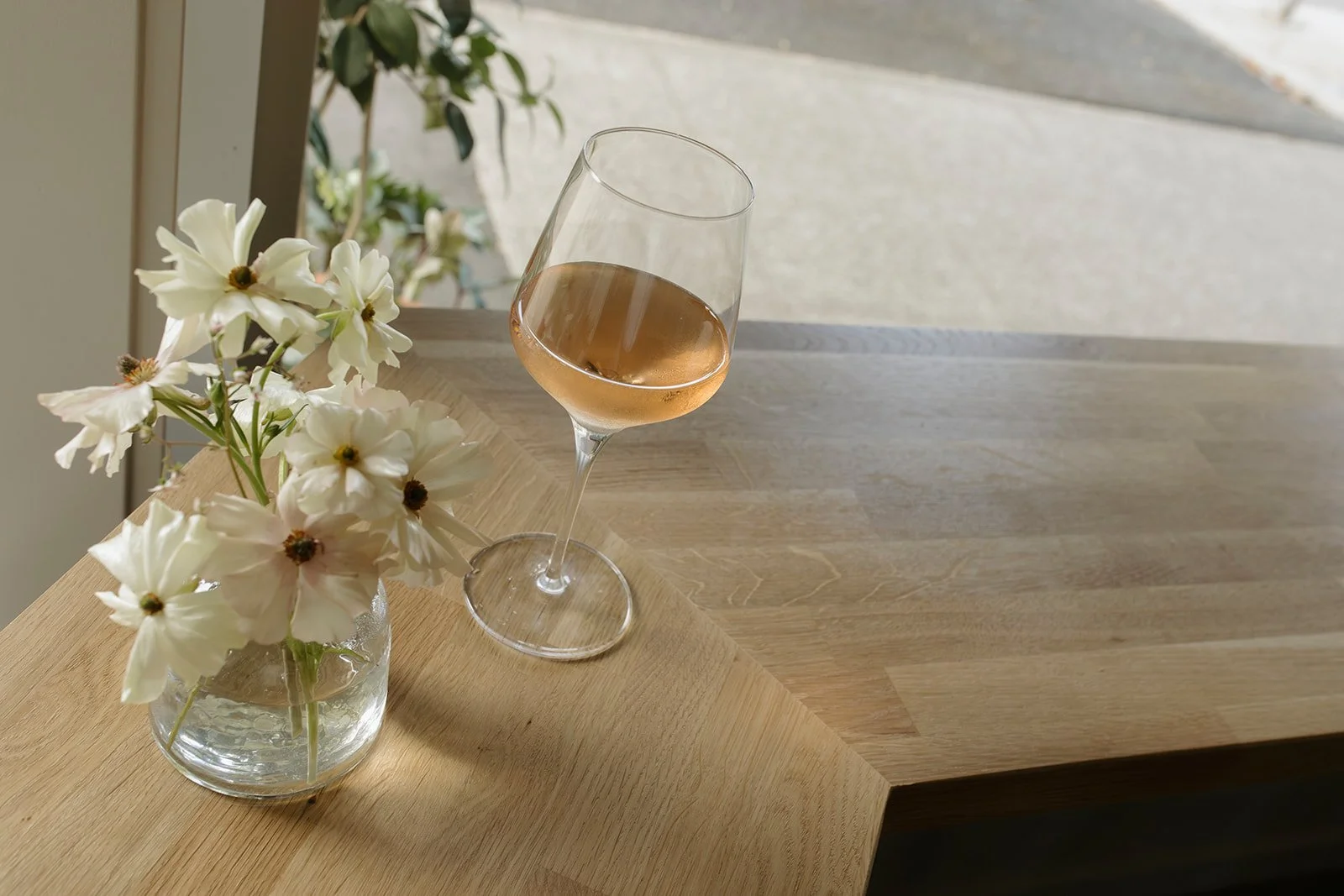 A glass of rosé wine on a wooden table near a vase with white flowers, next to a window.