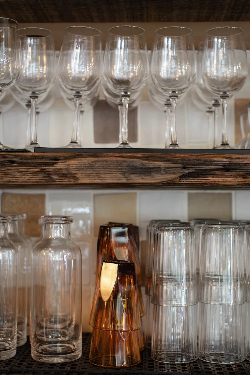 A collection of empty wine glasses on a wooden shelf and drinking glasses on a lower shelf in a restaurant or bar.