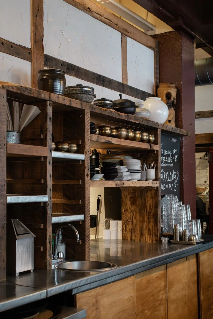 Wooden shelves with black pots, white and black bowls, gold cups, utensils, and glasses on a restaurant counter.