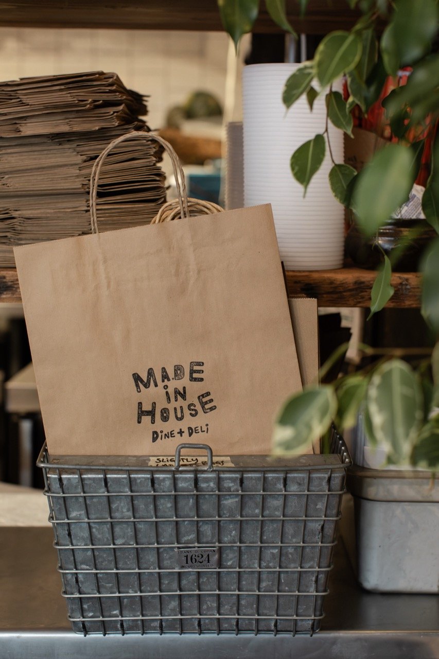A paper bag with 'Made in House Dine + Deli' written on it, placed in a metal basket. Behind and around the basket are stacks of paper bags, a white textured cup, and a leafy green plant. The setting appears to be a deli or cafe counter.