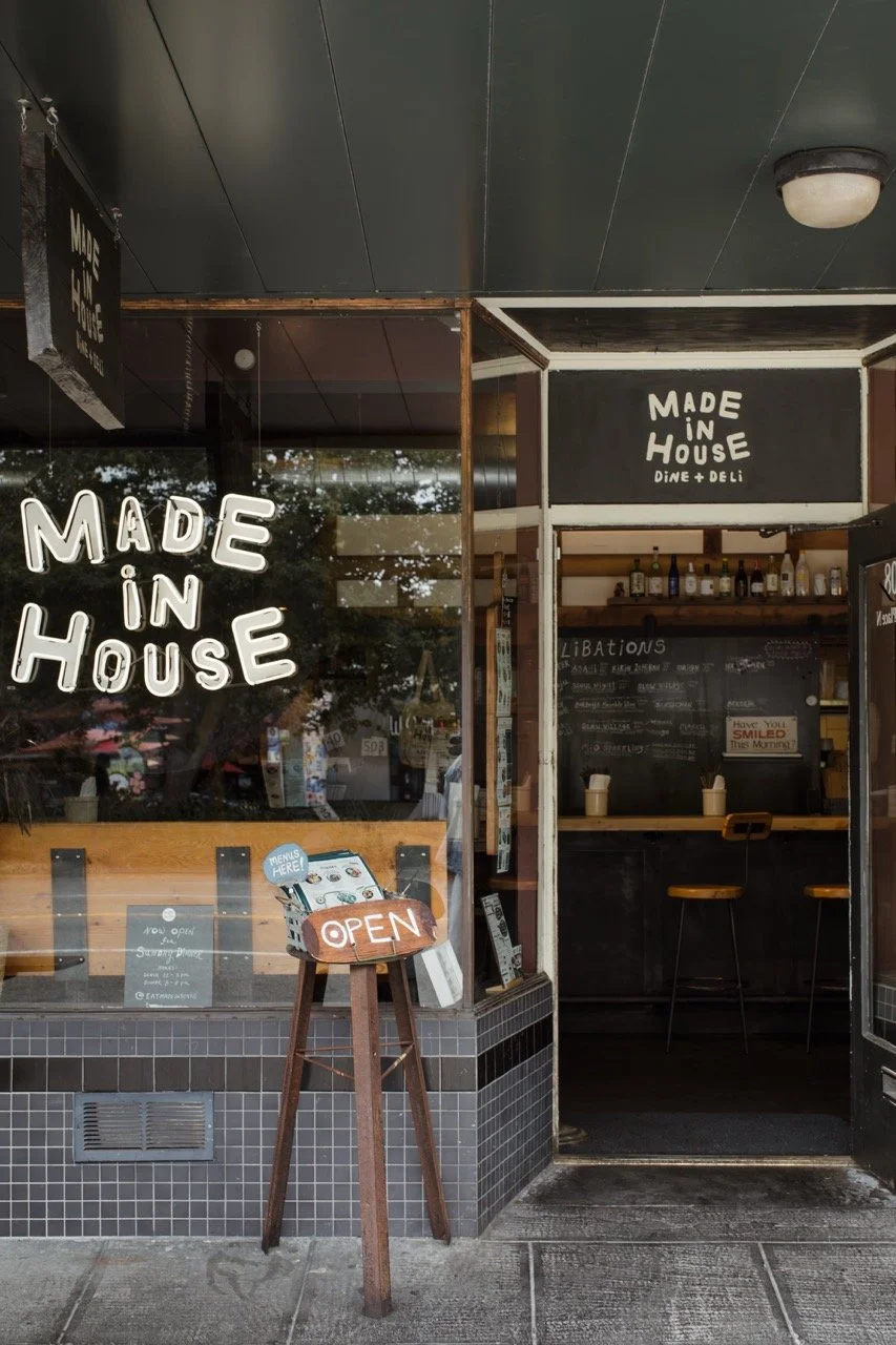 Exterior of a cafe with black and wood decor, displaying signs that read 'Made in House' and 'Open' on the window and door, with a menu board inside and bar stools visible.