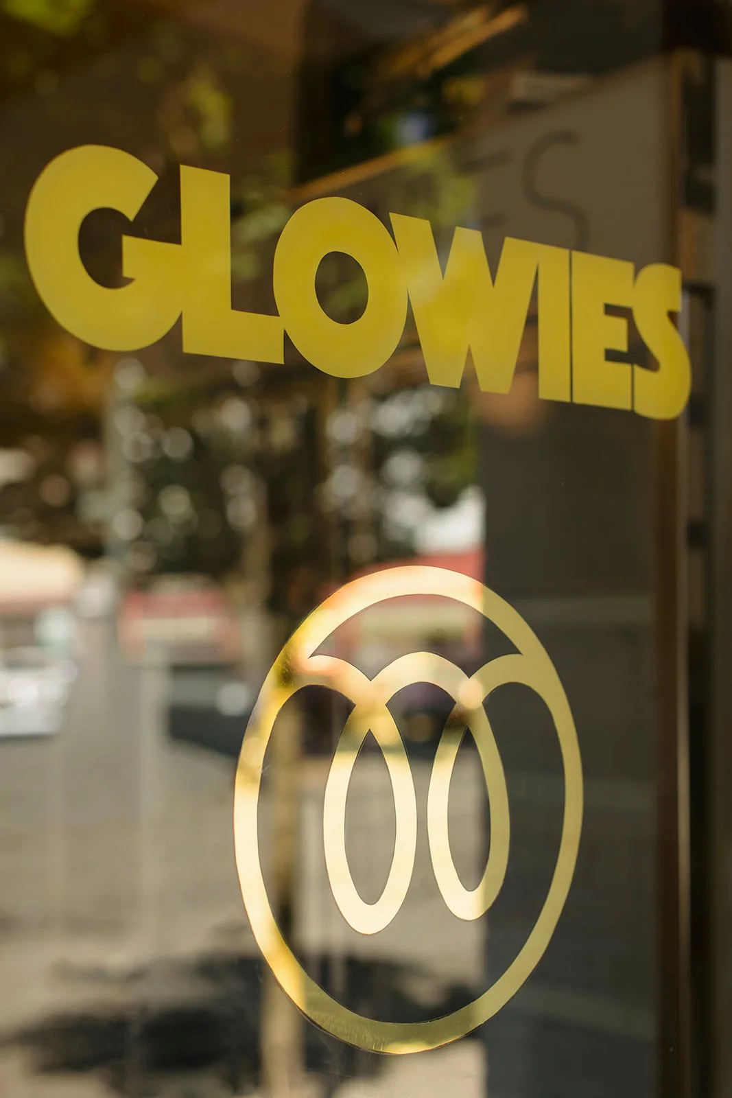 Yellow text on glass window reads 'GLOWS' with a smiley face logo below it. The reflection shows trees and a street scene outside.