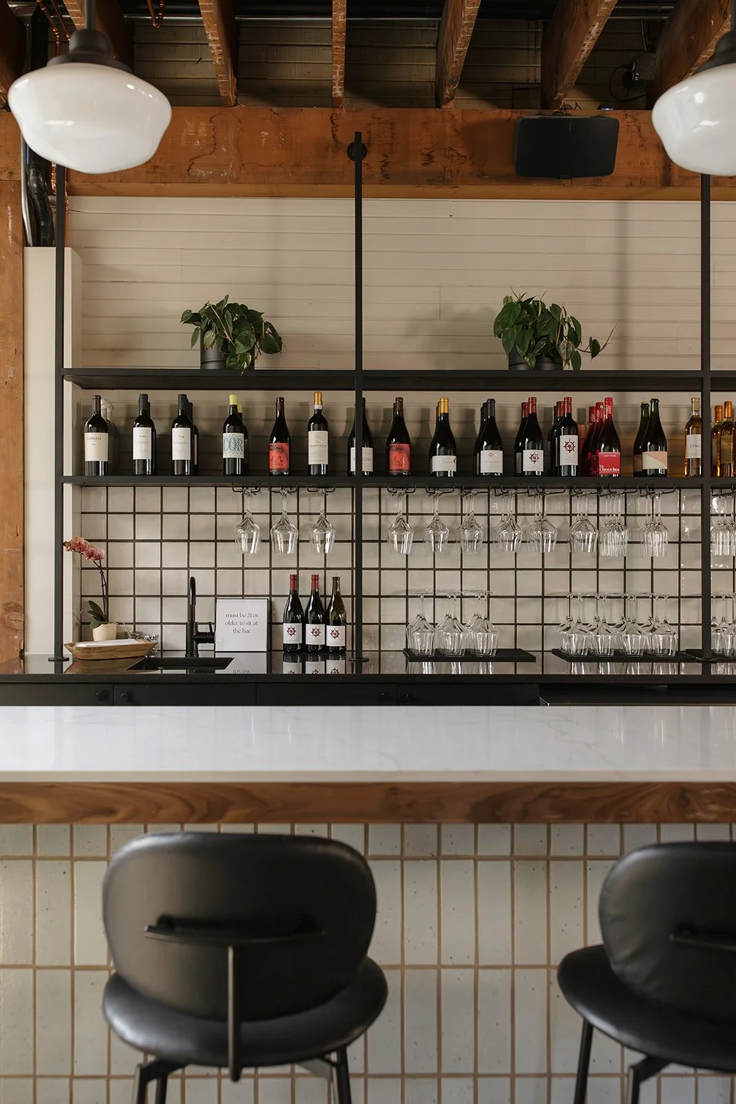 A modern bar with black metal shelves holding wine bottles, wine glasses, and potted plants. There is a white marble countertop and black bar stools in the foreground. The background includes a wood-beamed ceiling and a white paneled wall.