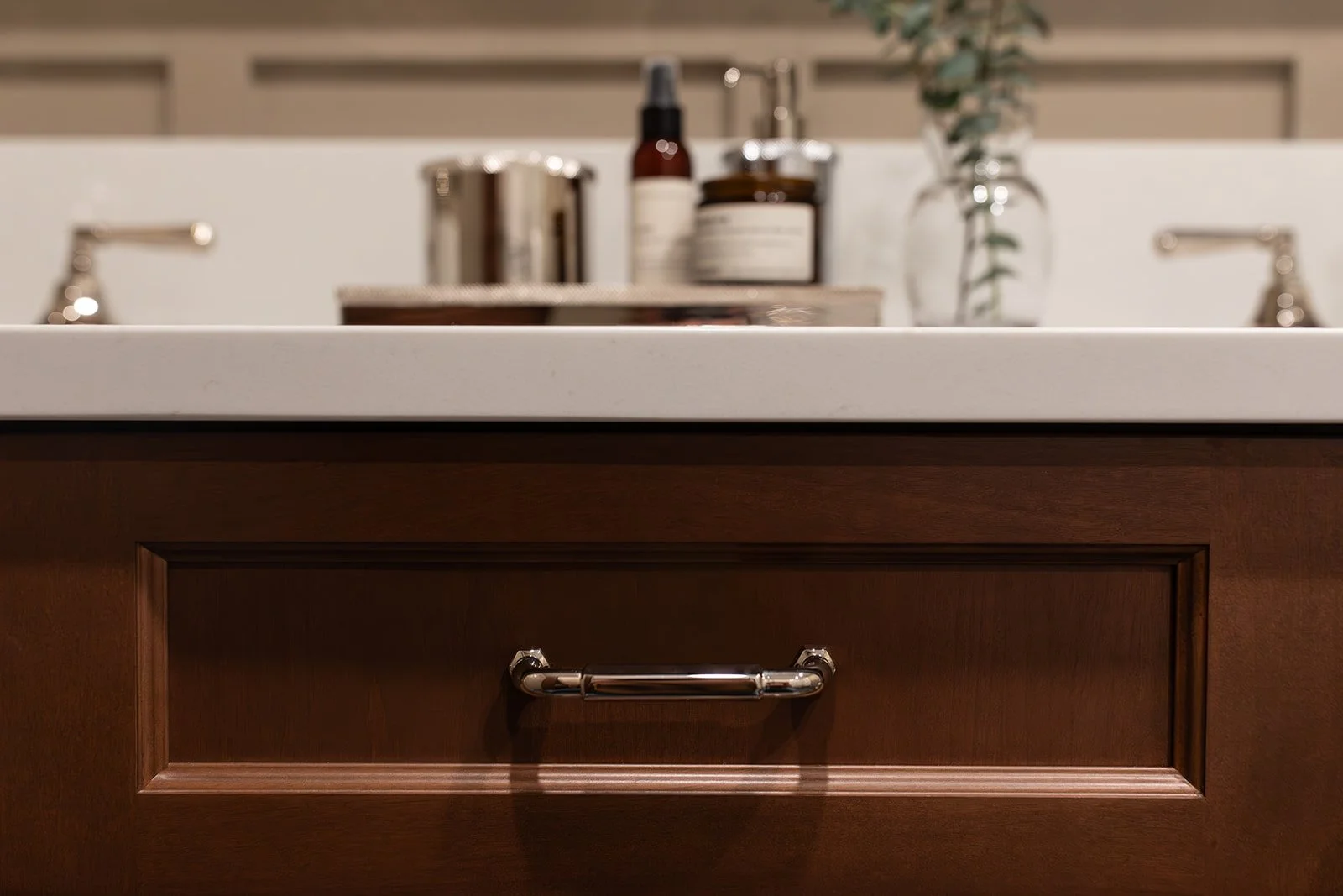Kitchen cabinet with a white countertop, wooden drawer with a silver handle, and blurred items on the countertop including bottles and a vase with greenery.