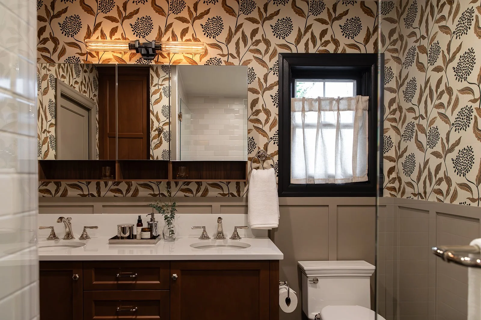 Bathroom with floral wallpaper, a mirror, a window with a curtain, a double sink with a tray of toiletries, a towel hanging on a ring, a toilet, and a part of a shower visible.