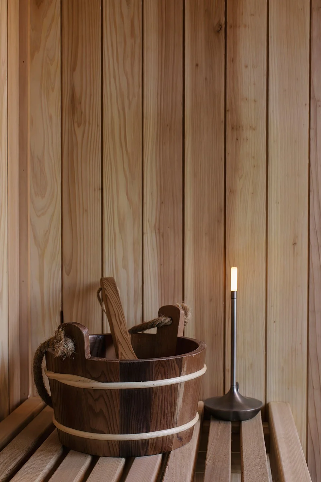 Wooden bucket with a wooden paddle inside, placed on a slatted wooden bench in a sauna room. A tall, modern lamp with a warm light is beside them.