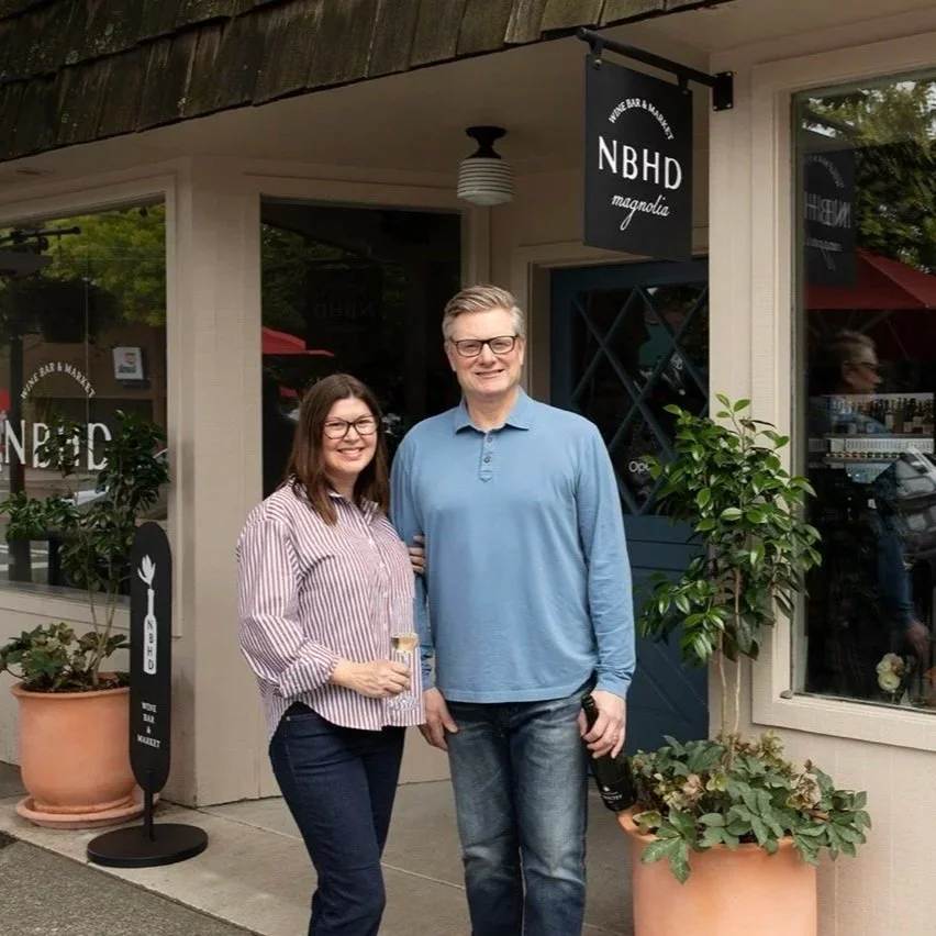 A smiling woman and man standing outside of a restaurant called NBHD, with potted plants and a sign, during the daytime.