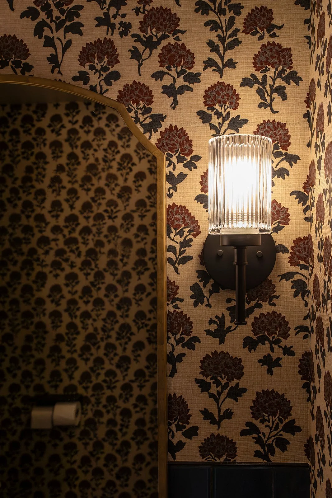 A wall with floral wallpaper featuring dark red and black flowers on a beige background, with a modern wall sconce light fixture with a ribbed glass shade and black base.