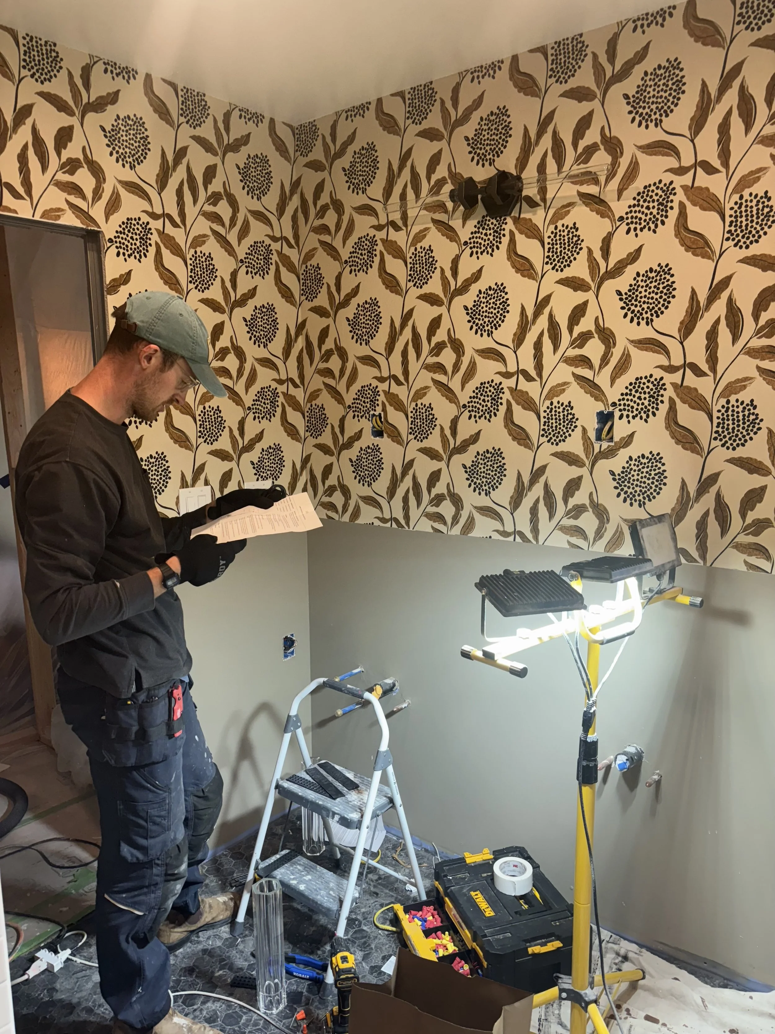 A man wearing a gray cap, safety glasses, and black gloves is working on a home renovation. He is standing in front of a partially finished wall with floral patterned wallpaper. There are construction tools, a step ladder, and electrical boxes around him, and a temporary work light illuminates the area.