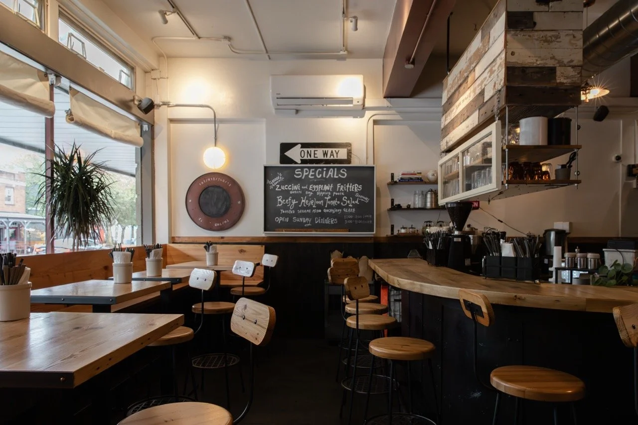 Interior of a cozy café with wooden tables and chairs, large windows, and a chalkboard menu.