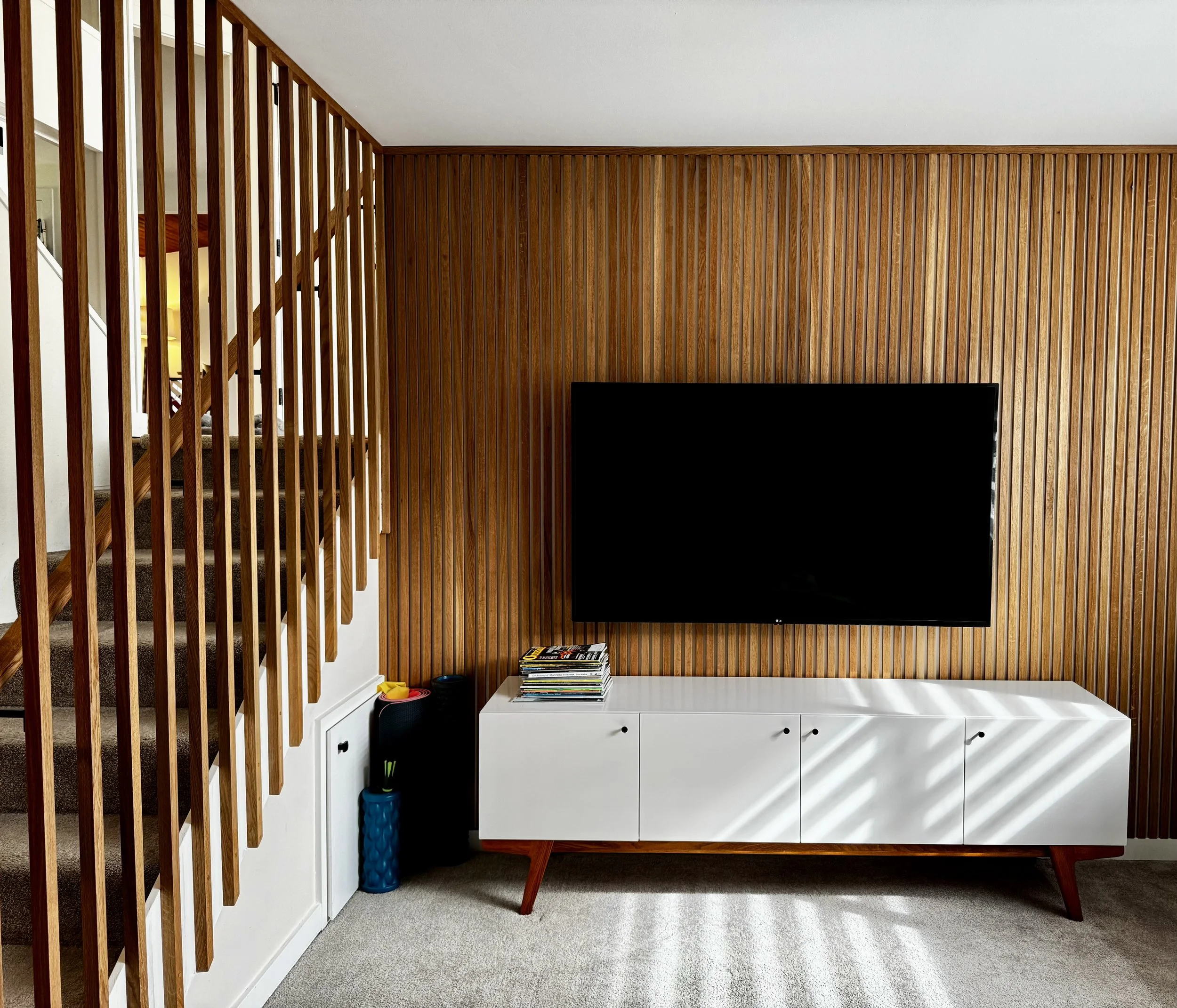 Living room with a wooden slat wall, a white cabinet, a black television, and a small pile of magazines on the cabinet. Part of a staircase with vertical wooden balusters is visible on the left. The room has neutral-colored carpet and a light-colored ceiling.