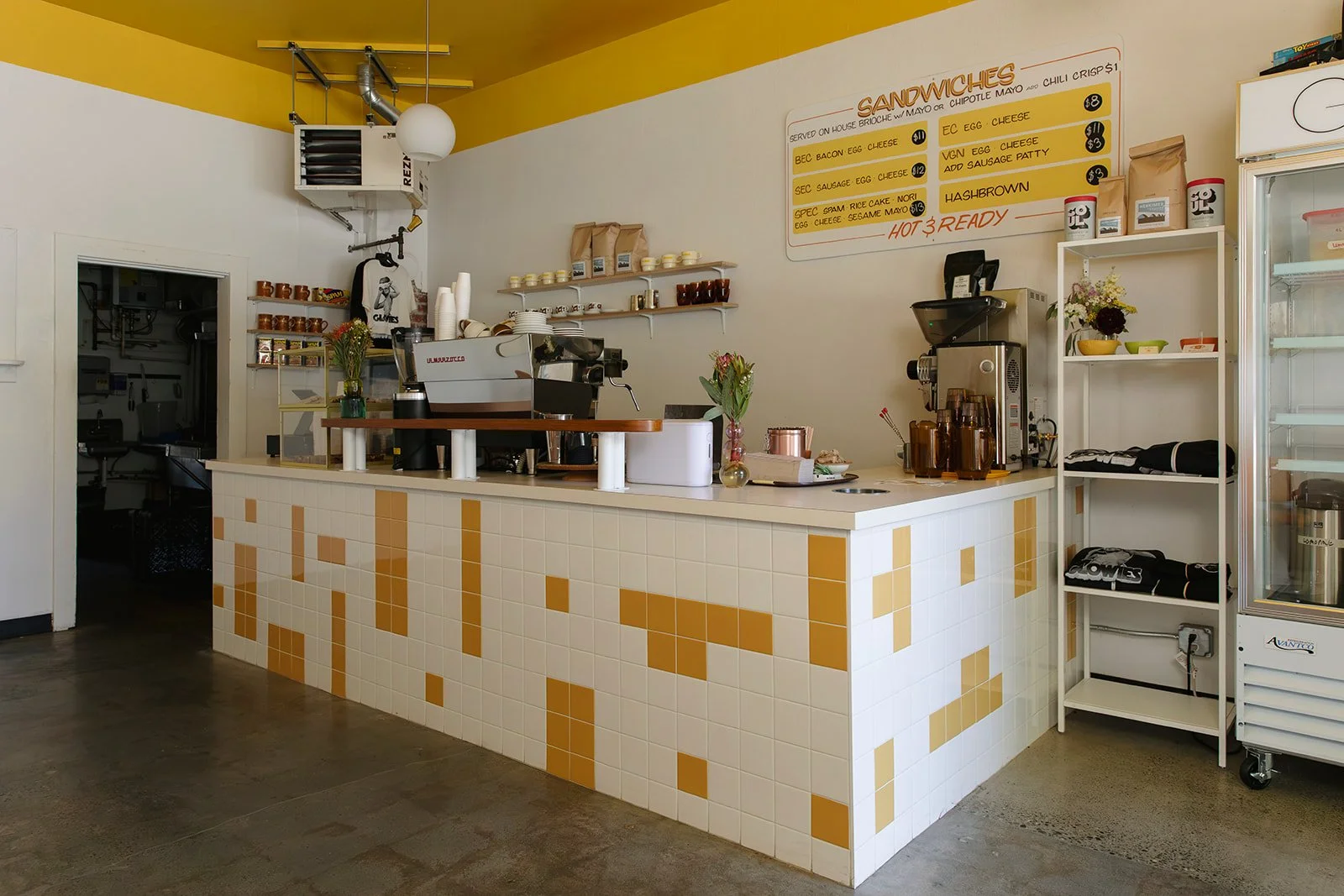 Counter area inside a small cafe or coffee shop with a white tiled front decorated with yellow tiles. Behind the counter, there is a coffee machine, a coffee grinder, and shelves with cups, jars, and napkins. On the wall, a menu board lists various s
