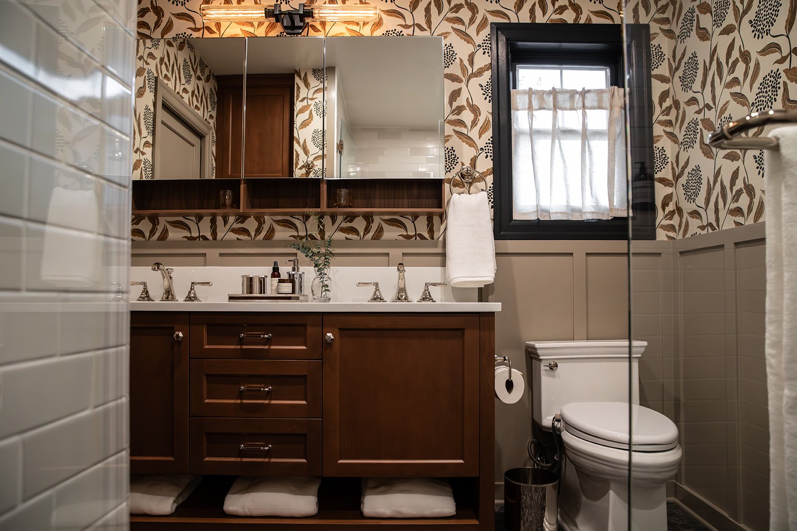 Bathroom with a wooden vanity, white countertop, mirror, window with curtain, floral wallpaper, and toilet.