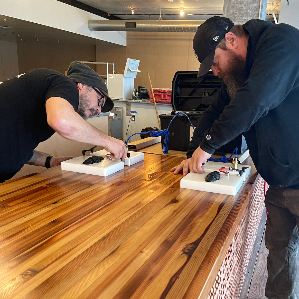 Two men working on electronic devices mounted on white foam bases at a wooden table in an industrial-style room.
