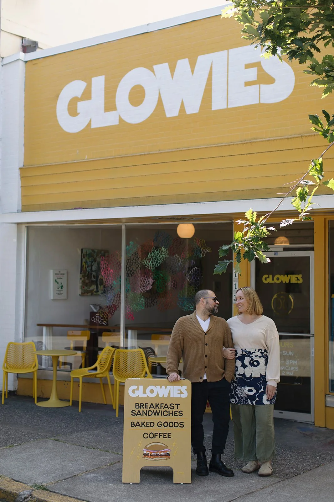 Two people, a man and woman, standing outside a yellow storefront called 'GLOWIES'. The man has a beard and glasses, wearing a brown cardigan, and the woman has red hair, wearing a cream sweater and a floral skirt. They are smiling at each other. A y