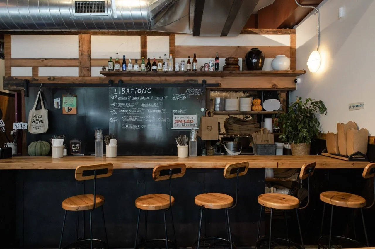 Coffee bar with bar stools, a chalkboard menu, shelves with bottles and dishes, and decorative indoor plants.