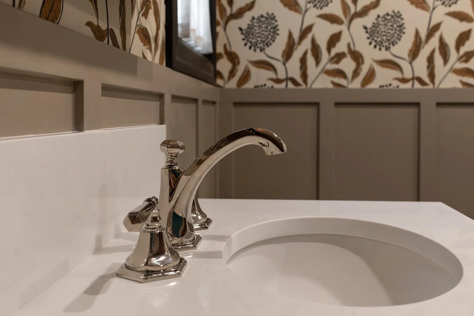 Close-up of a shiny silver bathroom sink faucet on a white countertop, with a decorative wallpaper featuring floral patterns in the background.