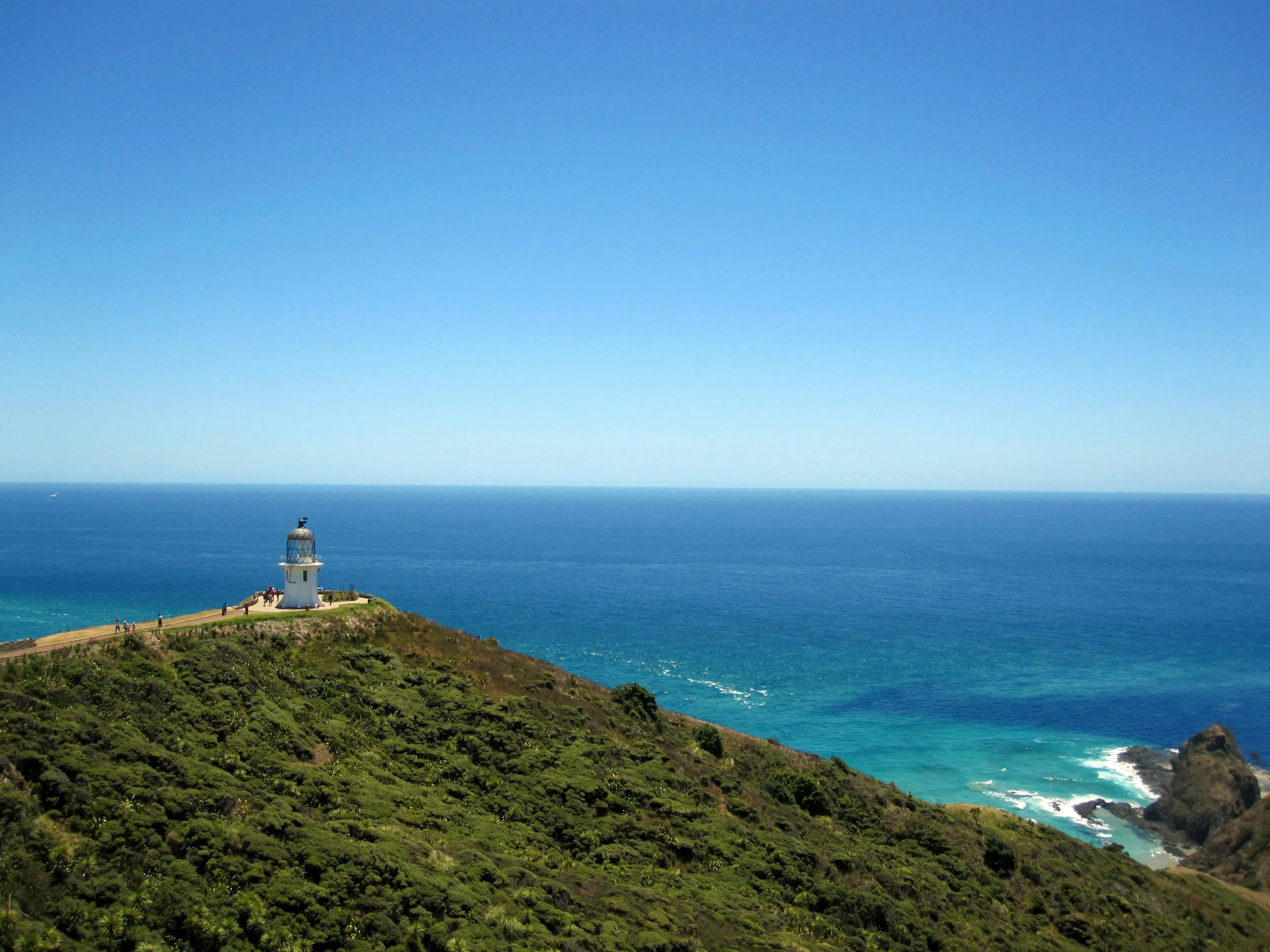 A coastal scene with a lighthouse on a green hillside, overlooking the ocean under a clear blue sky.