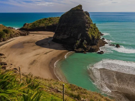 A large rock formation on a sandy beach with turquoise water and a cloudy sky.