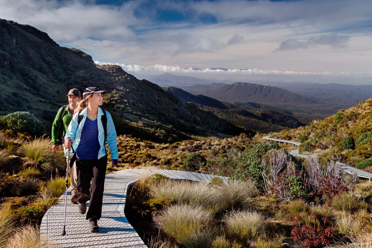Two women hiking on a winding trail in a mountainous landscape with hills, vegetation, and a cloudy sky in the background.