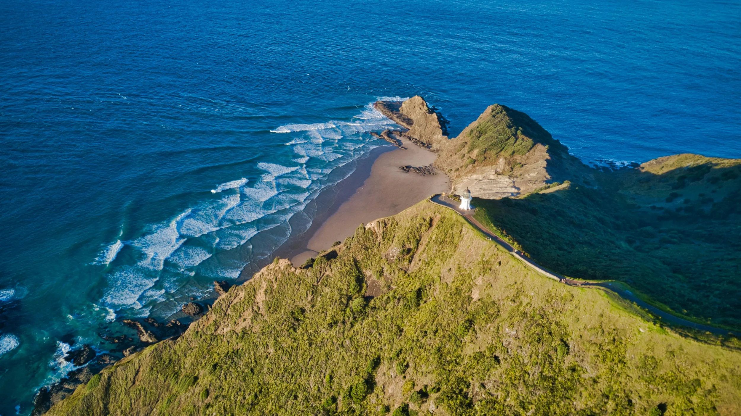 Aerial view of a rocky island with lush greenery, a lighthouse at the tip, surrounded by blue ocean water with visible waves.