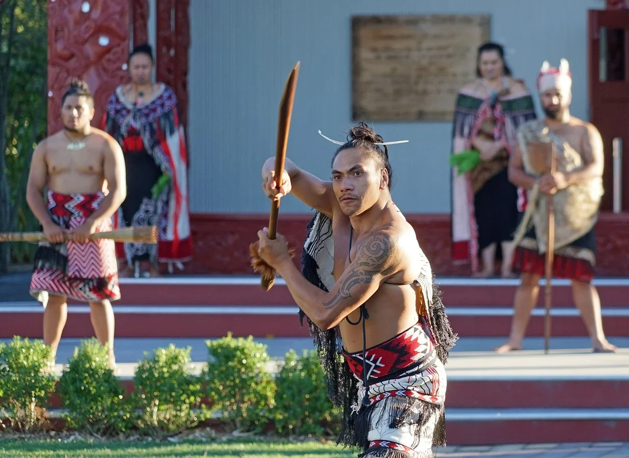 A native Maori warrior performing a traditional haka dance outdoors, holding a spear, with a group of performers in traditional attire standing behind him.