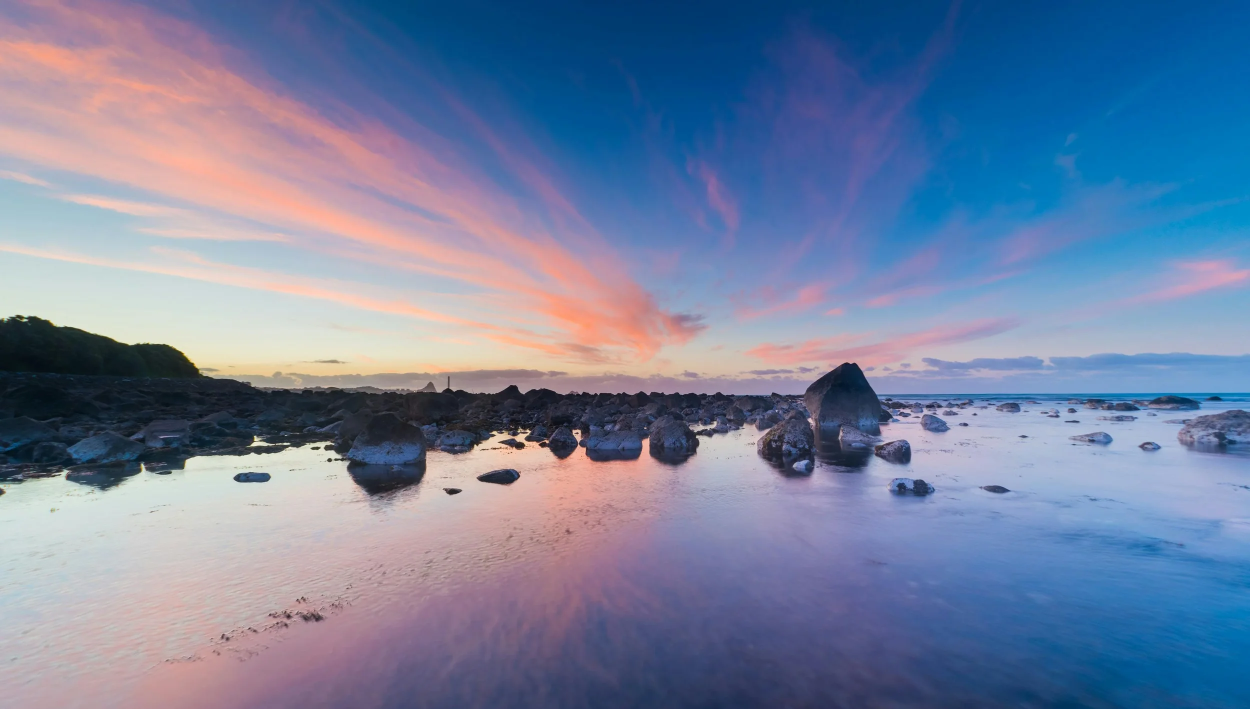 Sunset over a rocky shoreline with pink, purple, and blue sky reflections on calm water.